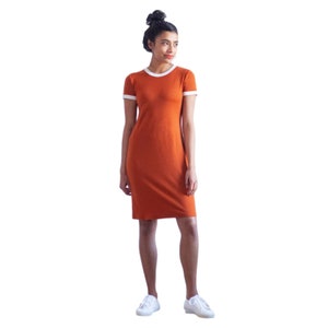 May include: A woman wearing a rust-colored short-sleeved dress with a white trim around the neckline and sleeves. She is wearing white sneakers.