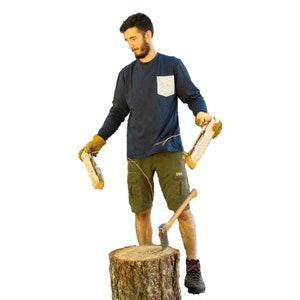 May include: A person wearing a dark blue long-sleeved shirt with a white pocket and green cargo shorts is holding two pieces of firewood. They are standing next to a tree stump with an axe leaning against it.