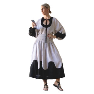 May include: A woman wearing a beige linen dress with a black and white wavy hemline. The dress has a black neckline with a circle cutout. She is holding a smartphone in her right hand.