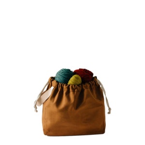 May include: A brown canvas drawstring bag with a white drawstring and a white handle. The bag is filled with balls of yarn in red, green, and yellow.