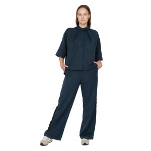 May include: A woman wearing a dark blue shirt and matching pants with black side stripes. The shirt has a collar and button closure.