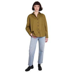 May include: An olive green button-down shirt with a single chest pocket, paired with light blue jeans. The outfit is completed with black shoes. The model is wearing a casual, everyday outfit.