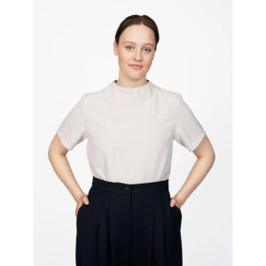 May include: A woman wearing a light pink, short-sleeved blouse with a high neckline and a pair of dark blue, high-waisted pants.