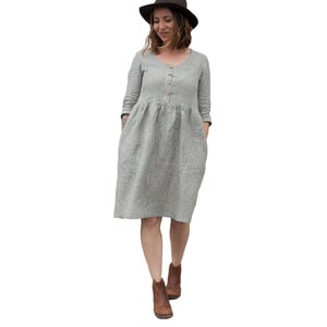 May include: A gray and white striped linen dress with a button-up front and 3/4 sleeves. The dress has a loose fit and a slightly gathered waist. The model is wearing brown ankle boots.