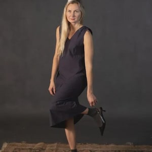 May include: A woman wearing a dark navy blue sleeveless dress with a V-neck and a party hat. She is wearing black ankle boots with a small heel. The background is a dark gray.