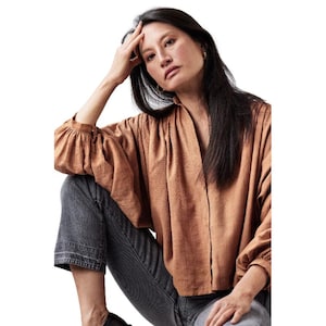 May include: A long-sleeved, loose-fitting blouse in a warm, muted copper-brown tone with a subtle textured pattern, featuring a V-neckline and gathered cuffs, paired with dark charcoal gray denim jeans that have a visible horizontal hem stitch.