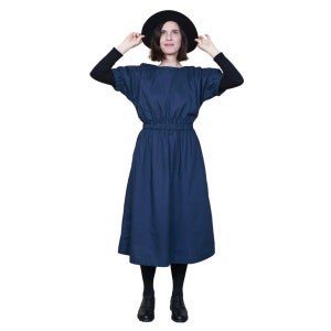 May include: A woman wearing a dark blue midi dress with a gathered waist and black short-sleeved shirt underneath. She is wearing a black hat and black shoes.