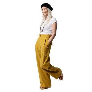May include: A woman wearing a white t-shirt, mustard yellow pants, a black beret, and a beaded necklace. She is standing with her hand in her pocket and looking to the side.
