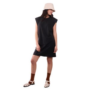 May include: A woman wearing a black sleeveless dress and a beige bucket hat. She is also wearing brown socks and white sandals. The dress is knee-length and has a simple, minimalist design. The outfit is set against a white background.