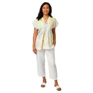 May include: A short-sleeved V-neck blouse featuring a light yellow, blue, and lavender floral pattern with vertical pleats at the neckline, paired with white cropped trousers and simple white slide sandals on a plain white background.