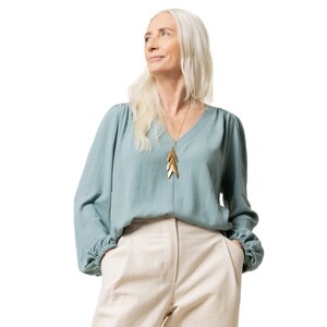 May include: A woman with long, white hair wearing a light blue blouse with a gold necklace and beige pants. She is looking up and to the right.