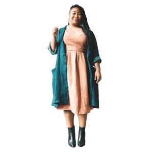 May include: A woman wearing a teal blue long-sleeved cardigan over a peach-colored midi dress. She is wearing black ankle boots and has long dark hair.