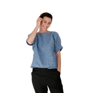 May include: A woman wearing a light blue linen top with rolled up sleeves and black pants. She is smiling and touching her hair.