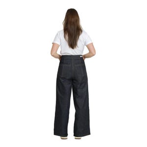 May include: Dark wash denim wide-leg pants with high waist and visible stitching. The pants are paired with a white t-shirt. The model is viewed from the back, showcasing the design and fit of the jeans. The pants are a classic style.