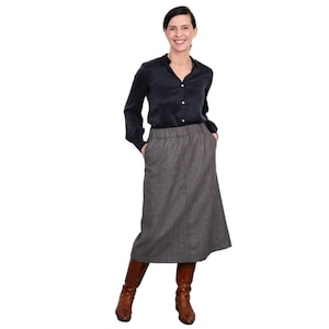 May include: A woman wearing a black long-sleeved shirt and a brown and black plaid skirt. She is wearing brown boots and has her hands in her pockets.