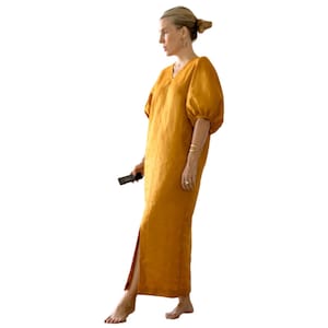 May include: A woman wearing a long, mustard yellow linen dress with a V-neck and puffed sleeves. The dress has a side slit and the woman is barefoot.