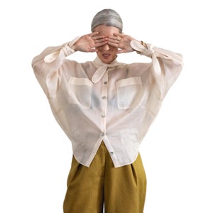 May include: A cream-colored, button-down blouse with a tie-neck detail and two front pockets. The sleeves are long and tied at the wrists. Paired with olive-green, high-waisted pants.