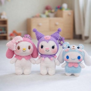 May include: Three handmade crochet dolls. The dolls are in the shape of cartoon characters. One doll is pink with a bow, one is purple, and the last is blue. Each doll has a keychain attached.