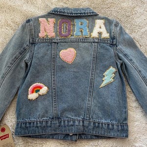 May include: A blue denim jacket with the name "NORA" spelled out in colorful felt letters. The jacket has several patches, including a rainbow, a heart, and a lightning bolt.