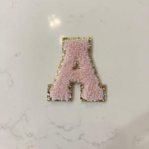 May include: Pink felt letter A with gold glitter trim. The letter is a decorative patch or applique.