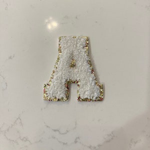 May include: White felt letter A with gold glitter trim. The letter is a puffy, raised design.