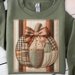 Può includere: Una felpa girocollo color verde oliva con una stampa rettangolare centrale raffigurante una zucca trapuntata. La zucca è composta da vari ritagli di tessuto nelle tonalità del marrone, beige e verde salvia, sormontata da un fiocco marrone.