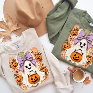 May include: A cream-coloured sweatshirt and an olive green sweatshirt, each featuring a graphic of a white ghost with a purple bow holding a jack-o'-lantern, surrounded by small pumpkins and flowers. A tan hat and a mug are also visible.