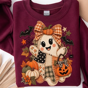 Puede incluir: Una sudadera color granate con un diseño de aplique central de un fantasma con un lazo a cuadros, sosteniendo un cubo de calabaza. El fantasma está rodeado de calabazas, hojas de otoño, murciélagos y una telaraña. La etiqueta Gildan es visible.