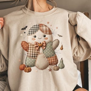 Peut inclure: Un sweat-shirt à col rond beige avec un motif central représentant un bonhomme en pain d'épice en patchwork. La figurine porte un nœud papillon à carreaux et est entourée de petites citrouilles, chauves-souris, toiles d'araignée et étoiles.