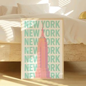 Puede incluir: Una impresión enmarcada con una ilustración rosa y naranja del Empire State Building en la ciudad de Nueva York. El fondo es blanco con el texto "NEW YORK" repetido en color turquesa.