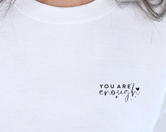 Op de afbeelding: Witte longsleeve trui met de tekst "YOU ARE enough" in zwarte letters met een klein hartje.