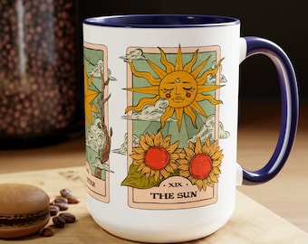 May include: White ceramic mug with a blue rim and handle. The mug features a tarot card design with a sun and sunflowers. The card reads "XIX. THE SUN".