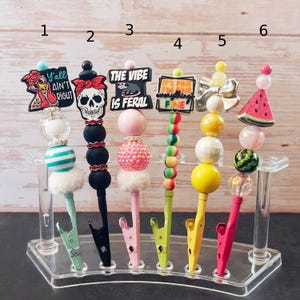 May include: A collection of six decorative pen clips with various designs. The clips feature charms with text like "Y'all Ain't Right" and "The Vibe Is Feral." Other charms include a skull, bow, and watermelon. Each clip has a unique color and bead arrangement.