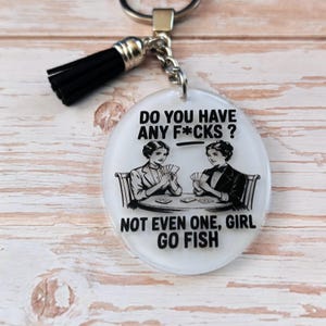 May include: Oval keychain with a black tassel. The white pendant features a black and white illustration of two women playing cards, with the text "DO YOU HAVE ANY F*CKS? NOT EVEN ONE, GIRL GO FISH."