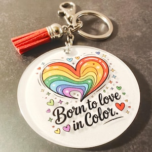 May include: A round keychain with a rainbow heart design and the text "Born to love in Color." The keychain has a red tassel and a silver-colored clasp.