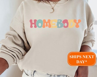May include: A beige sweatshirt with the word "HOMEBODY" printed in colorful, rounded letters. The letters are in shades of pink, yellow, blue, and green.