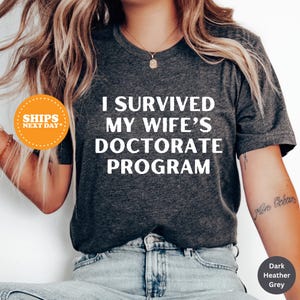 May include: A dark heather grey t-shirt with the text "I survived my wife's doctorate program" printed in white.