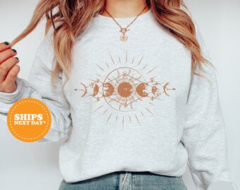 May include: A gray sweatshirt with a brown sun and moon phases design. The design features a sun with rays and five moons in different phases. The sweatshirt is shown on a model wearing blue jeans.