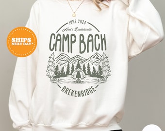 May include: White sweatshirt with a green graphic design of mountains, trees, and a tent. The text "JUNE 2024 Ava's Bachelorette CAMP BACH BRECKENRIDGE" is printed on the sweatshirt.