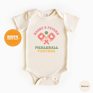May include: A cream-colored baby onesie with the text "Daddy's Future Pickleball Partner" in a retro font. The text is surrounded by two pink pickleball paddles with a green ball in the center.