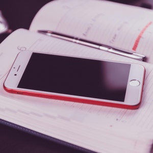 Può includere: Uno smartphone bianco con cornice rossa è appoggiato su un'agenda aperta. Una penna d'argento è posata sulle pagine dell'agenda. L'agenda è aperta su una pagina con note manoscritte e il numero 6.