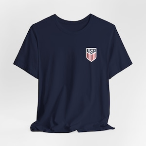 Pode incluir: Camiseta azul marinho com um pequeno emblema dos EUA no peito. O emblema apresenta um design de escudo com listras vermelhas e brancas e as letras "USA" em branco.