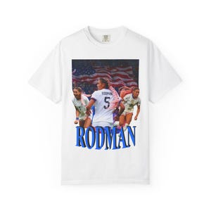 May include: White t-shirt with a graphic of a soccer player and the name "RODMAN" in blue. The player wears a white jersey with the number 5. The background features the American flag.