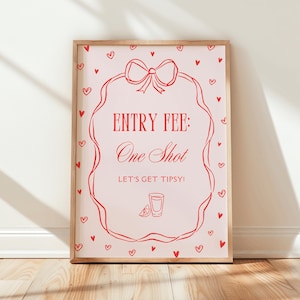 May include: A framed print with a light pink background and red text and illustrations. The text reads "ENTRY FEE: One Shot. Let's get tipsy!" with a bow and hearts. The frame is made of light-colored wood.