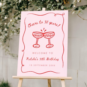 May include: A pink sign on a wooden easel with red text and illustrations. The sign reads "Cheers to 30 years!" and "Welcome to Natalie's 30th Birthday, 18 September 20XX" with two champagne glasses.
