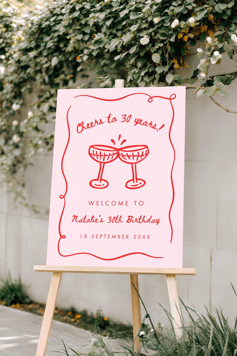 May include: A pink sign on a wooden easel with red text and illustrations. The sign reads "Cheers to 30 years!" and "Welcome to Natalie's 30th Birthday, 18 September 20XX" with two champagne glasses.