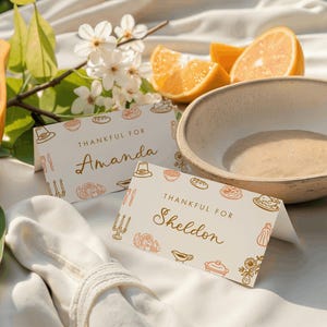 May include: A still-life image featuring two place cards that read "Thankful For Amanda" and "Thankful For Sheldon". The cards are decorated with Thanksgiving-themed illustrations. Sliced papaya, orange wedges, and a ceramic bowl are also present.