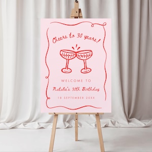 May include: A pink sign on a wooden easel with red text and illustrations. The sign reads "Cheers to 30 years!" and "Welcome to Natalie's 30th Birthday" with the date "18 September 20XX". Two red champagne glasses are illustrated.