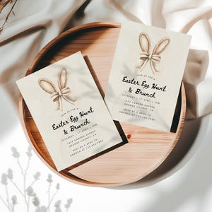 May include: Two cream-colored cards with bunny ears and a bow on the top. The text on the cards reads "Join us for a Easter Egg Hunt & Brunch" with the date and time of the event. The cards are on a wooden tray.