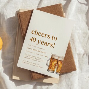 May include: A cream-colored invitation with the text "cheers to 40 years!" in brown lettering. The invitation includes details for John's 40th birthday, with watercolor illustrations of beer mugs. The invitation rests on a stack of books.